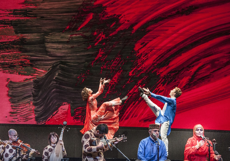 Two dancers lift their legs high into the air. Musicians are in the foreground while an abstract black and red mural hangs behind them.